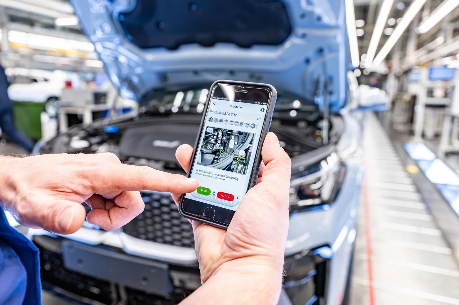Successful AI implementations involve understanding how the people utilizing the model already work, treating workflow design as seriously as model design. BMW did this by integrating a new model into an app that factory inspectors already carried. Here we see a hand holding a smartphone with an app in the foreground. The app has a green and red button at the bottom of the screen, which displays the layout of a car engine, with green circles and lines overlaid. In the background is a car assembly plant with a car’s front hood open, showing its engine.
